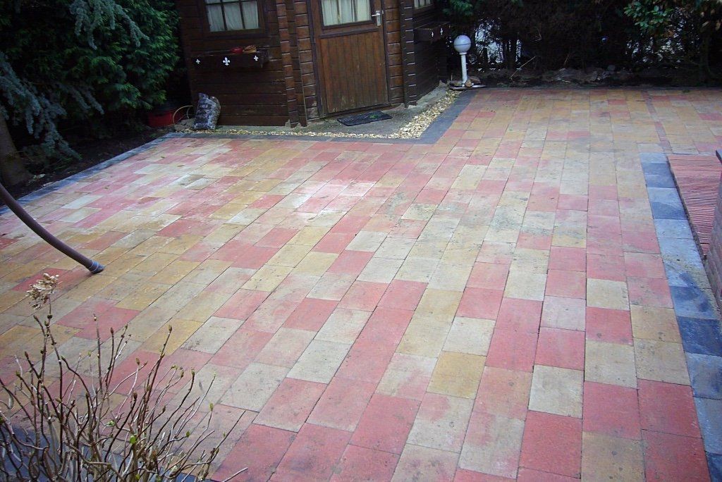 Een stenen terras met een houten schuur op de achtergrond, omgeven door planten en afgewerkt met donkere en lichte stenen.