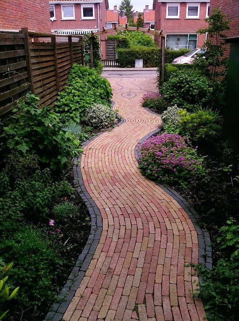 Een bakstenen pad slingert door een weelderige tuin, omzoomd met groen en bloemen, en leidt naar de huizen.