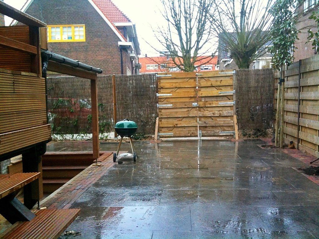 Een regenachtige achtertuin met een barbecue op een nat terras, omgeven door houten schuttingen en een houten constructie.