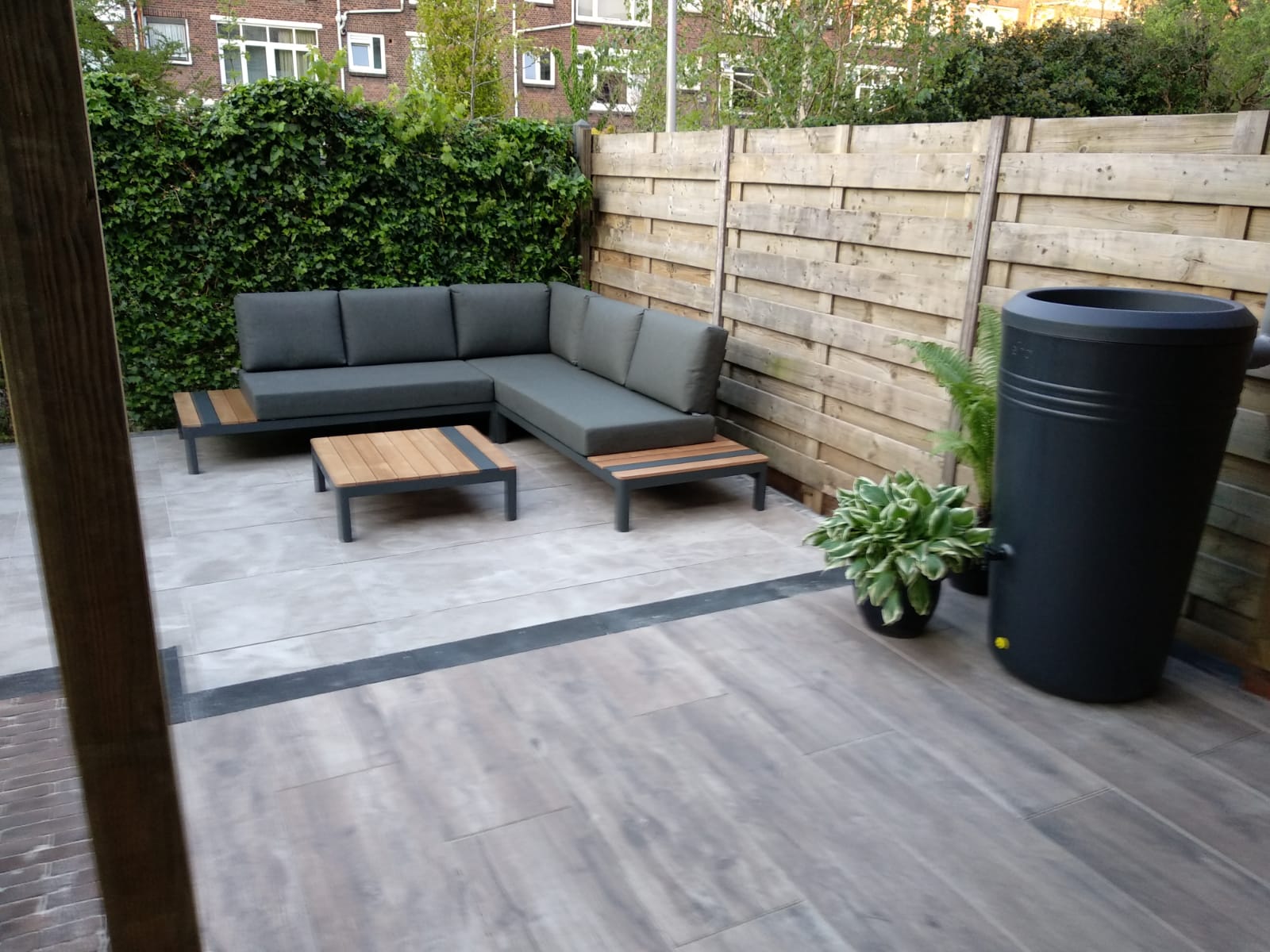 Terras met grijze hoekbank, houten salontafel en regenton. Groene muur en houten schutting op de achtergrond.