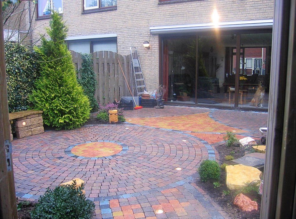 Een rondvormig terras van bakstenen, dat leidt naar glazen deuren van een huis. Groene begroeiing en een houten schutting omringen het gebied.