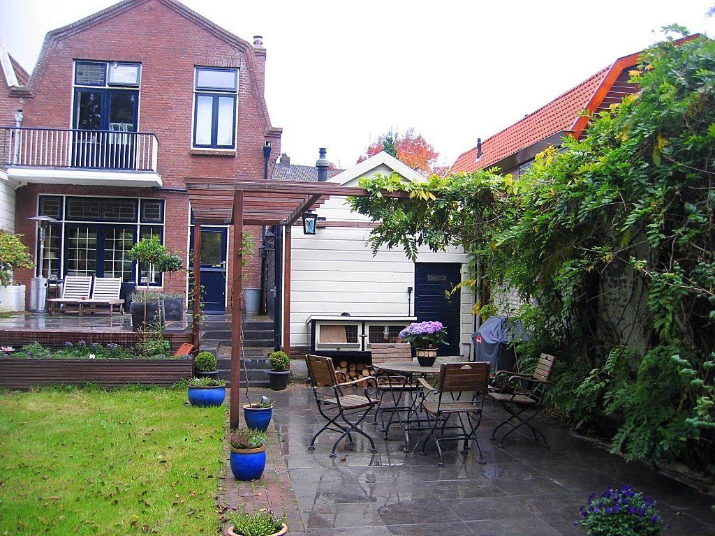 Achtertuin met bakstenen huis, terras en gazon. Houten tafel en stoelen onder een met klimplanten begroeide pergola.