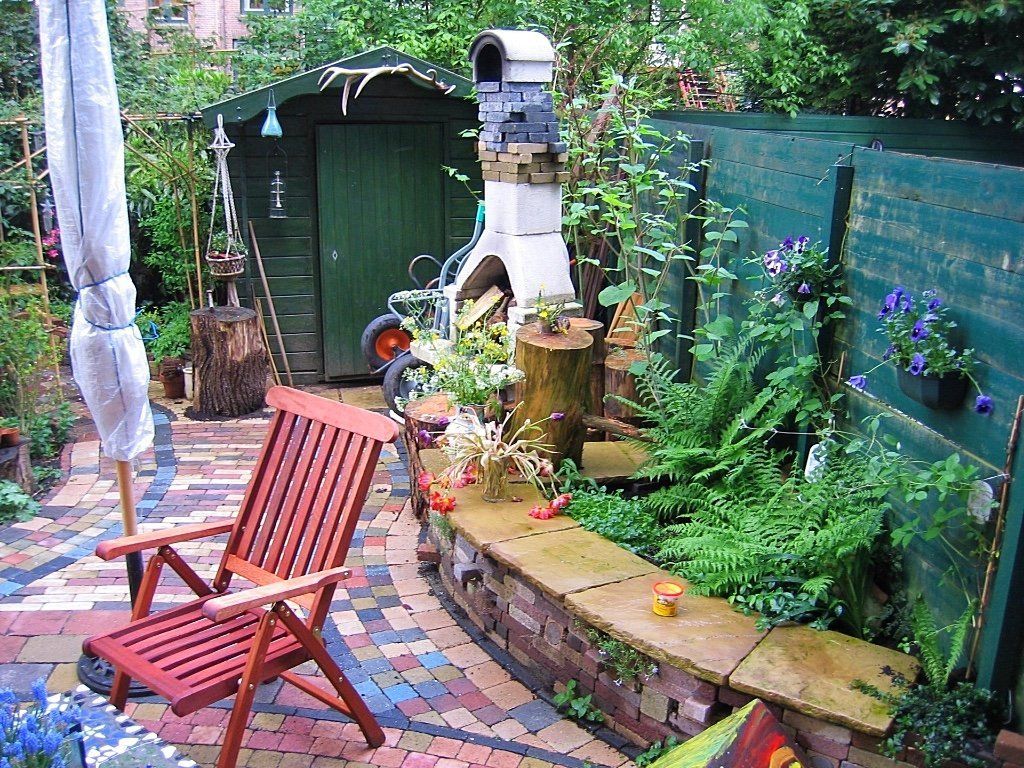 Gezellige tuinscène met bakstenen terras, houten stoel, bloembed met stenen randen, groene schuur en open haard.