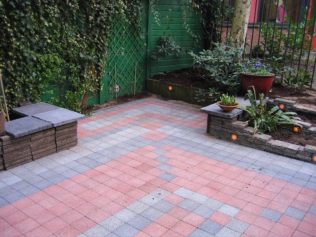 Een terras van bakstenen met rode, grijze en blauwe bestrating, omgeven door planten en een groene schutting.