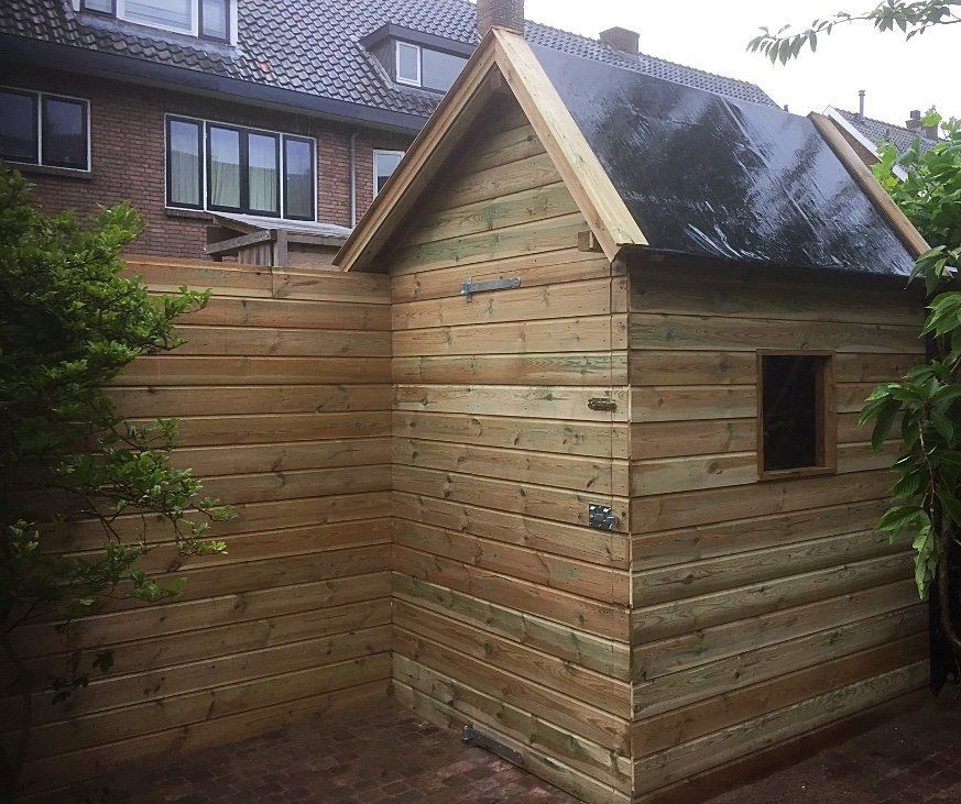 Houten schuur met klein raam en zwart zeildoekdak, gebouwd tegen een horizontale houten schutting.