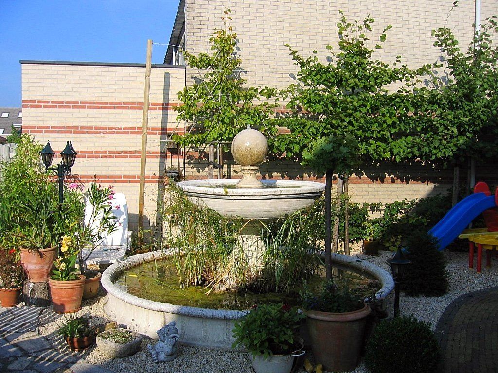 Fontein in een tuin met bomen, potplanten en een bakstenen gebouw op de achtergrond.