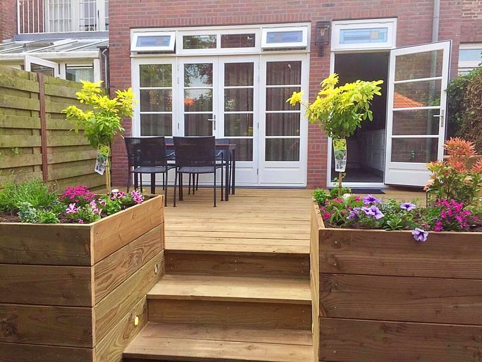 Houten terras met plantenbakken, een tafel en openslaande deuren naar een bakstenen gebouw.