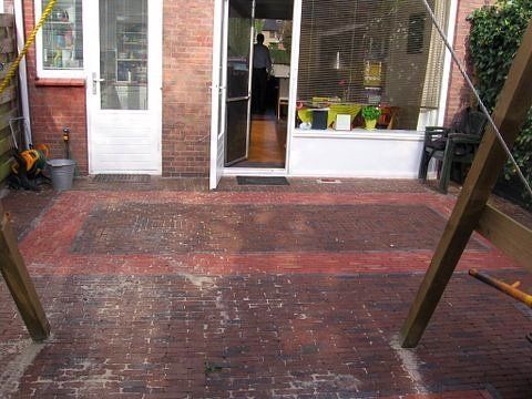Een stenen terras met rode rand, dat naar een open deur leidt. Rechts staan ​​houten palen voor een schommel.