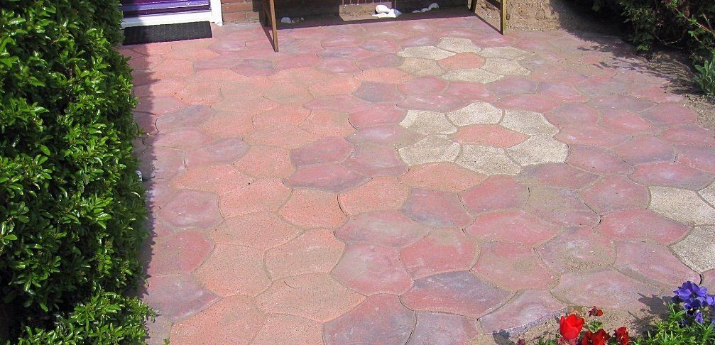 Terras van rode en beige stenen met bloemmotieven, omgeven door struiken en enkele bloemen.