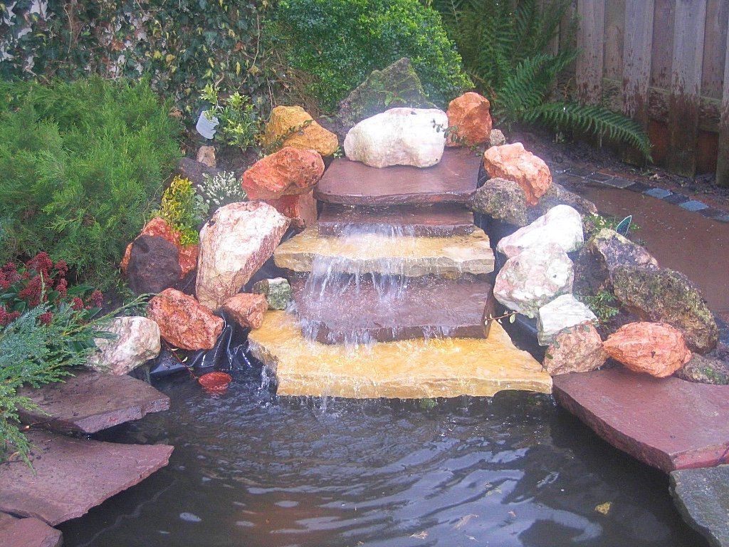 Waterfontein met een trapsgewijze stenen waterval die in een vijver stroomt, omgeven door kleurrijke rotsen en groen.