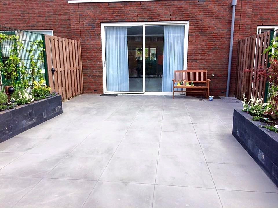 Terras met grijze tegelvloer, bakstenen muur, glazen schuifdeuren, houten bank en bloemperken.