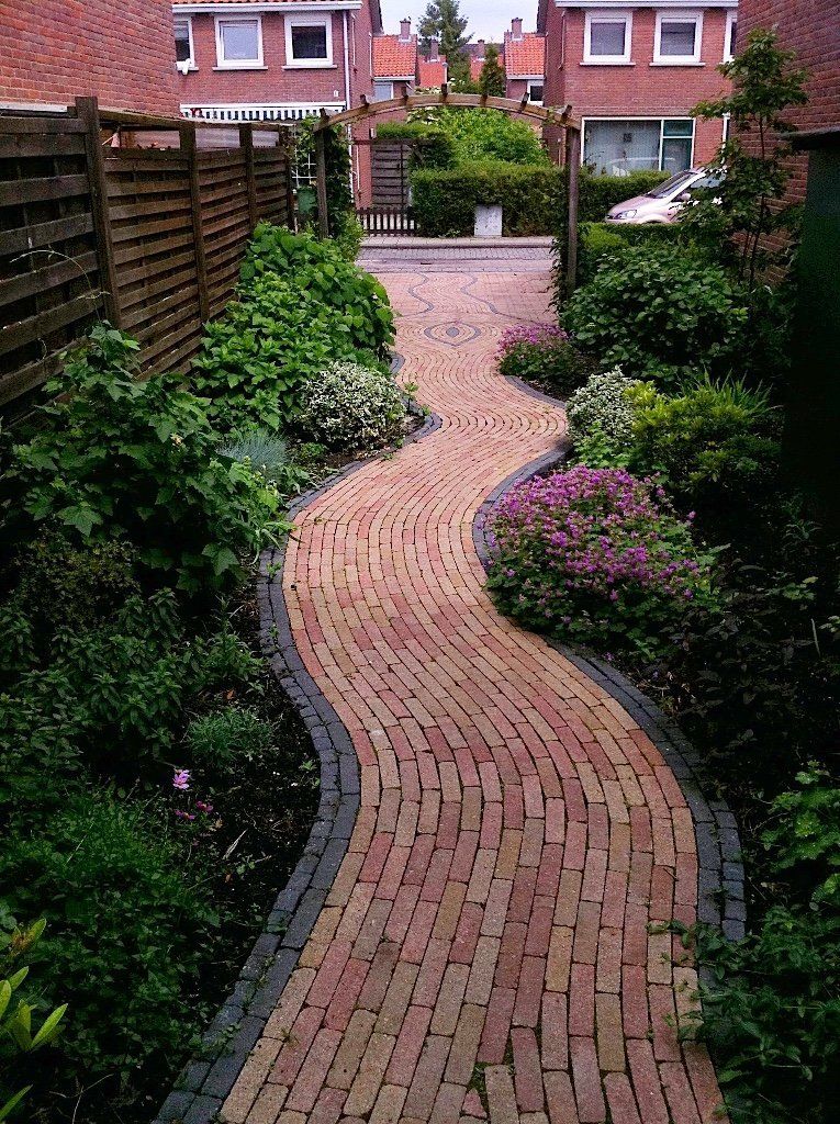 Een bakstenen pad kronkelt door een tuin, omzoomd met groene planten, richting woongebouwen.