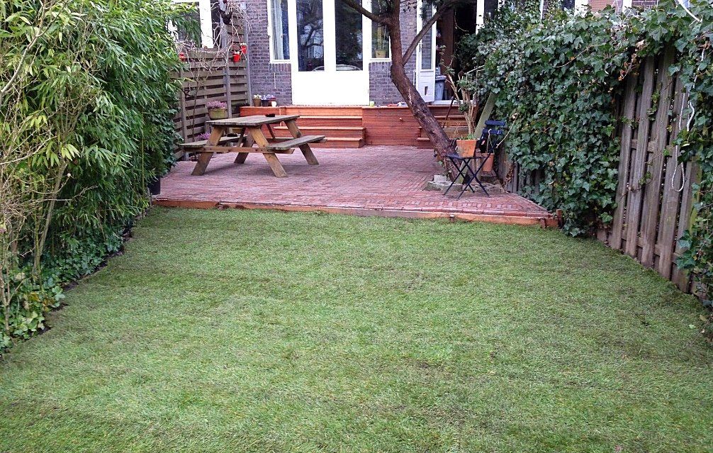 Een groen gazon leidt naar een terras met een picknicktafel, omgeven door hagen en hekken.