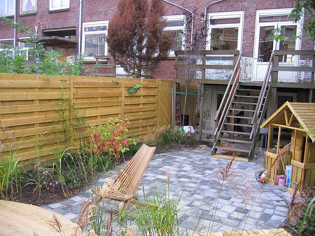 Achtertuin met houten schutting, terras, trap naar balkon en een speelhuisje.