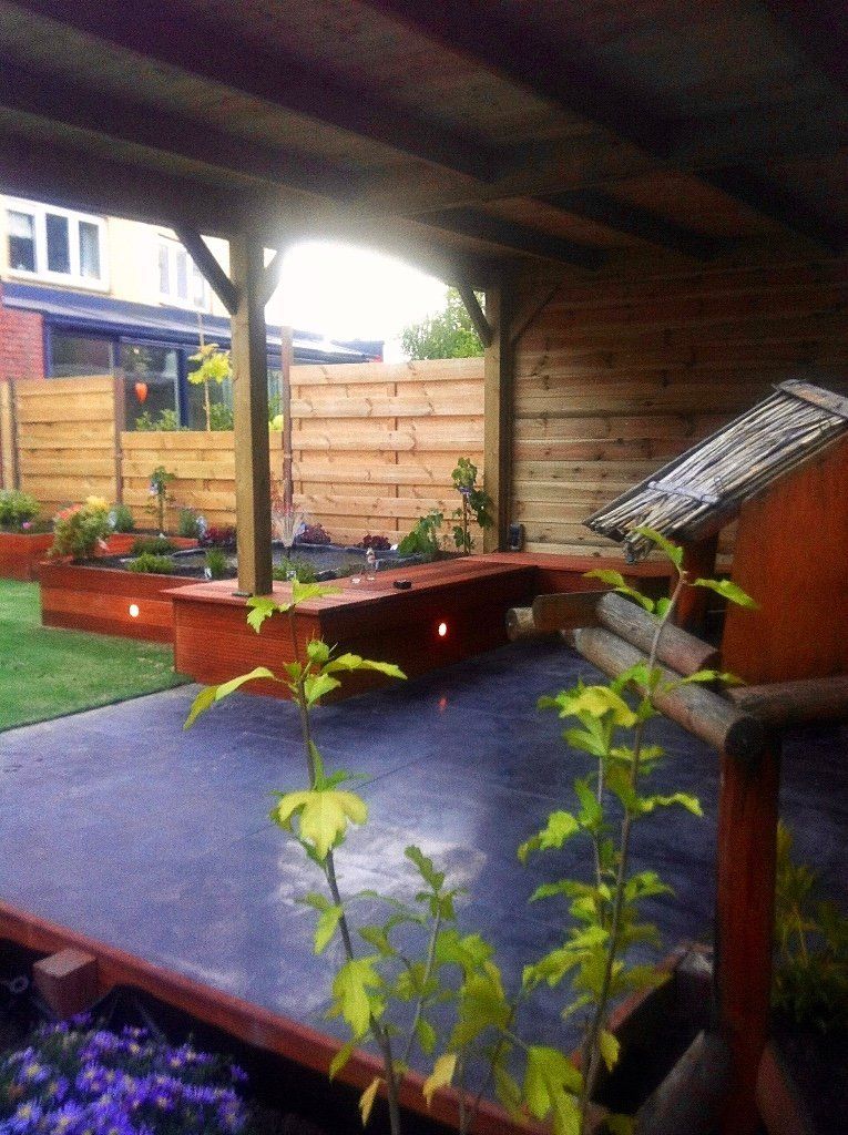 Een buitenruimte met houten frame, ingebouwde zitgelegenheid, plantenbakken en een overdekt gedeelte, en een groen gazon.