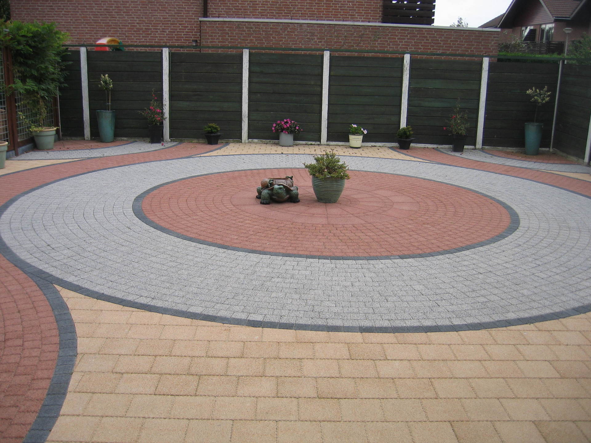 Rond terras, bestraat met bakstenen en natuursteen, kleine planten en een donkerhouten schutting.