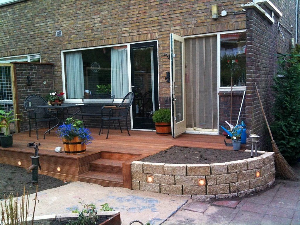 Houten terras met bakstenen huisje, kleine traptreden, bloembed en zitgelegenheid buiten.