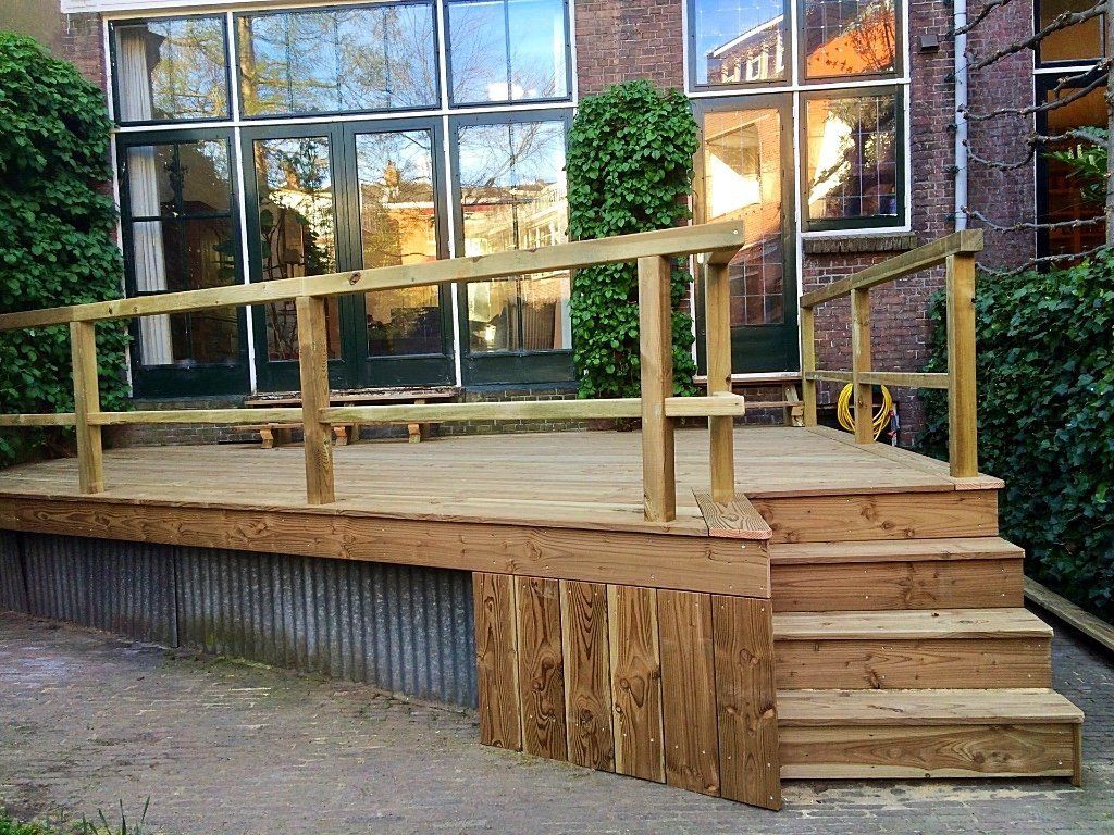 Houten terras met trap en balustrade, naast een gebouw met grote ramen.