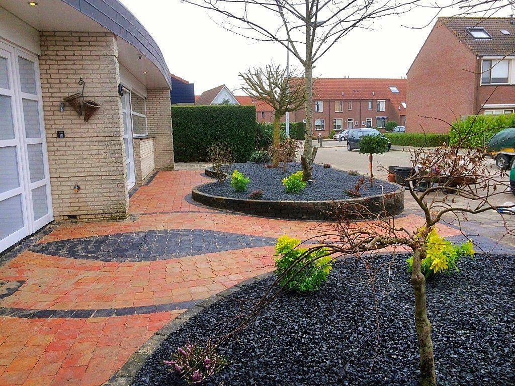 Tuinaanleg met bakstenen en zwarte natuursteen voor een huis. Er zijn kleine bomen en struiken geplant.