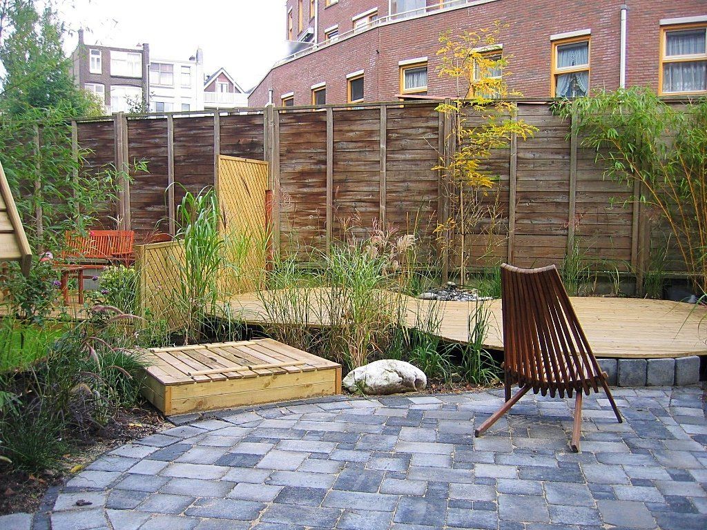 Terras in de achtertuin met houten tuinmeubelen, zandbak en schutting, met een bakstenen gebouw op de achtergrond.