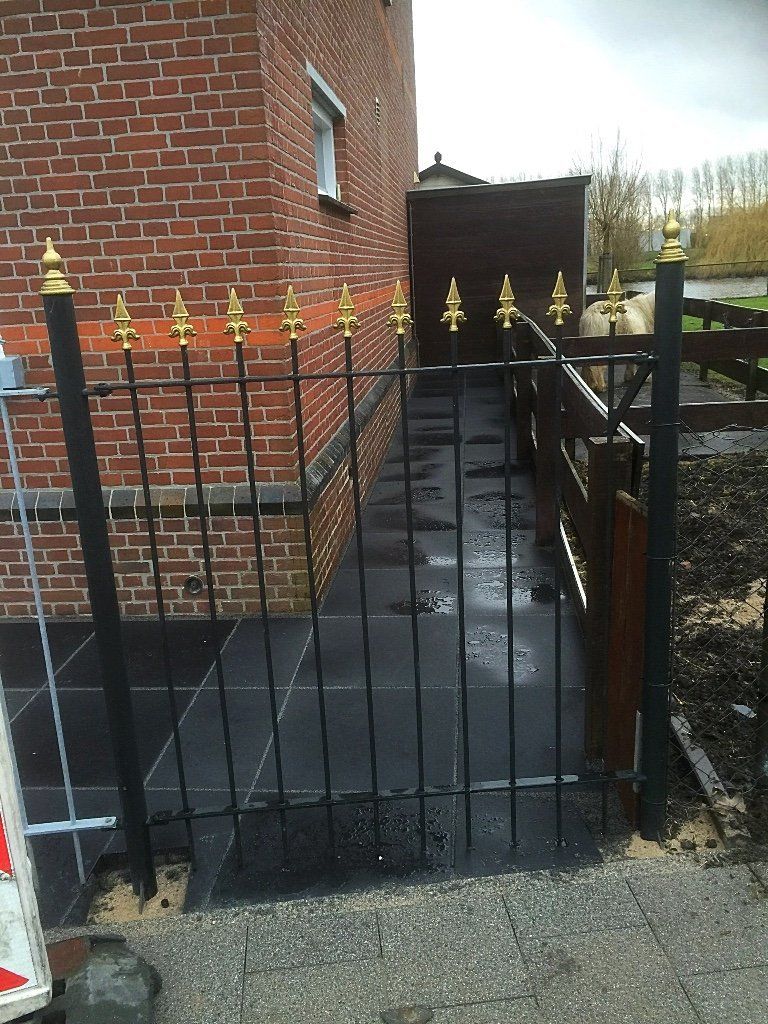 Zwarte metalen poort met gouden sierelementen, bakstenen gebouw en donkerstenen pad.