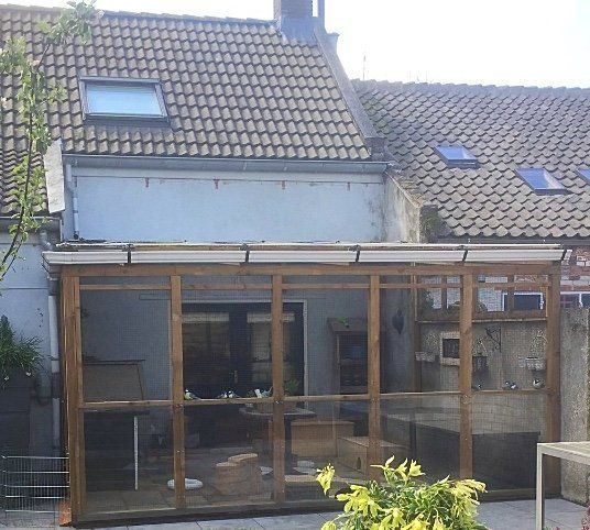 Een houten volière, aangebouwd aan de buitenkant van een gebouw, met een pannendak.