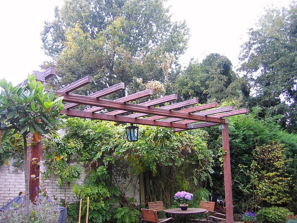 Een houten pergola in een tuin, begroeid met groen, met een tafel en stoelen en een hangende lantaarn.