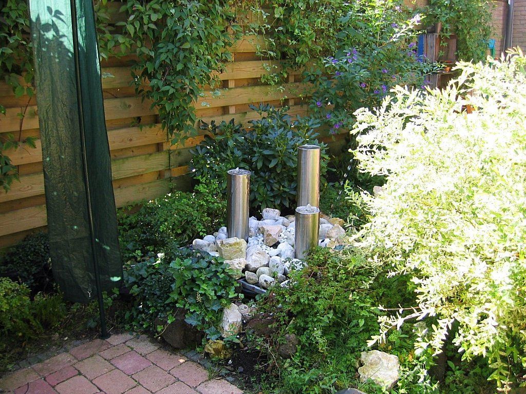 Tuin met een waterfontein, voorzien van roestvrijstalen pilaren, omgeven door rotsen en weelderig groen.