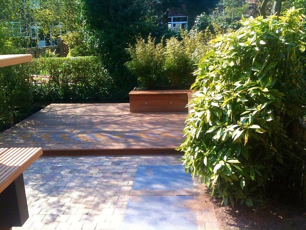 Houten terras met bank, omgeven door groen en een stenen patio.
