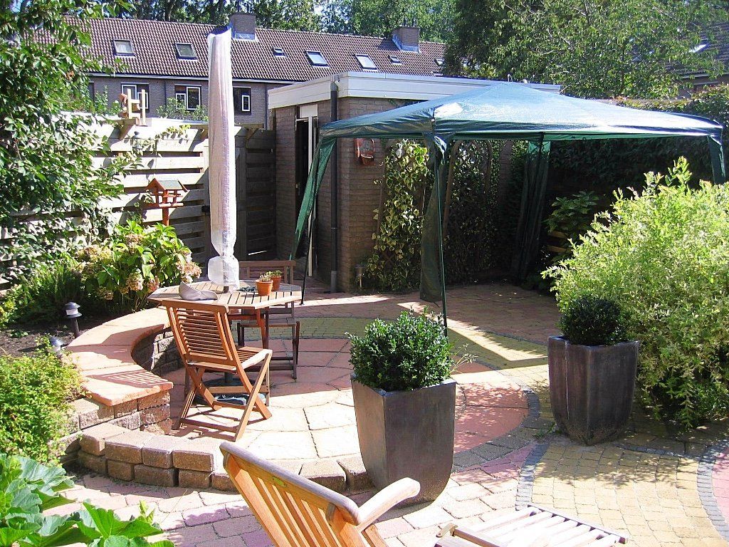 Terras met bakstenen muren, houten meubels en een overkapping. Tuin met groen en een gebouw op de achtergrond.