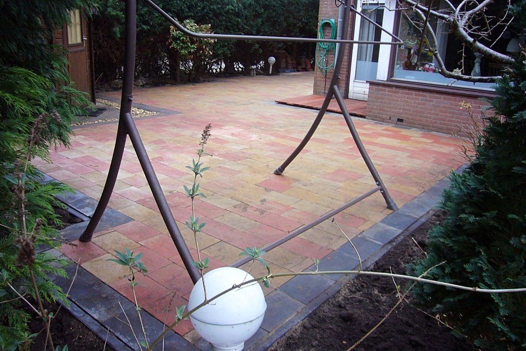 Terras bestraat met kleurrijke bakstenen, metalen schommel, omgeven door groen.