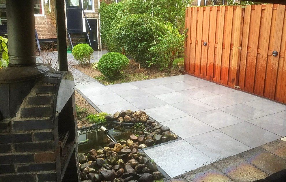 Terras met grijze natuurstenen tegels, houten schutting, kleine vijver en gesnoeide groene struiken.