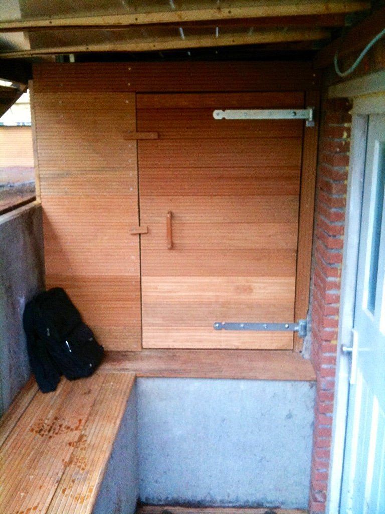 Houten deur en muur. Zitgedeelte. Metalen scharnieren en handgreep. Aangrenzende bakstenen muur met witte deur.