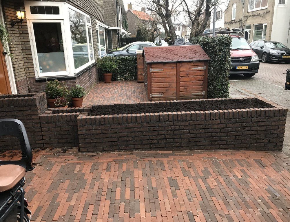 Bakstenen plantenbakken voor een huis, een rood bakstenen pad, een houten schuur en auto's geparkeerd op straat.