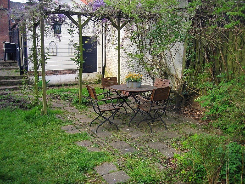 Een tuin met een tafel en stoelen onder een houten pergola, omgeven door groen en een bakstenen gebouw.