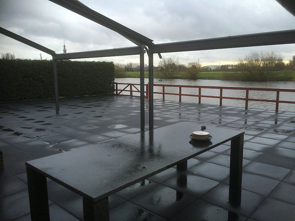 Terras met een tafel, nat van de regen. Bewolkte lucht en een waterpartij op de achtergrond.