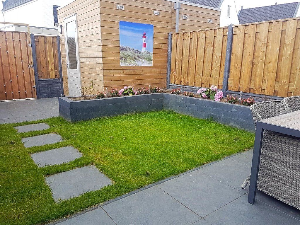 Kleine achtertuin met groen gras, een stenen terras en houten schuttingen. Aan de houten muur hangt een schilderij.