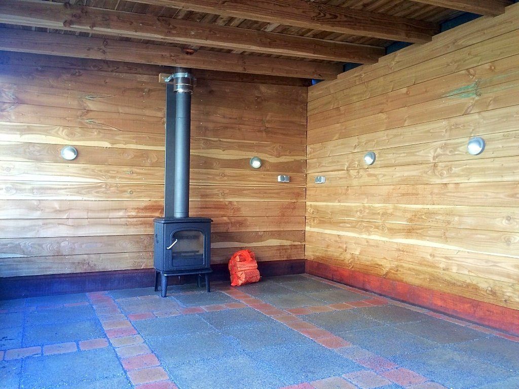 Een kamer met houten lambrisering, een houtkachel met schoorsteen, inbouwspots en een rode bakstenen rand op de vloer.