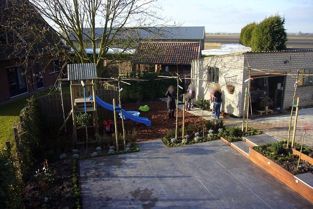 Achtertuin met speeltoestel, zitgedeelte voor volwassenen en een bijgebouw, met een betonnen gedeelte en plantenperken met bomen.