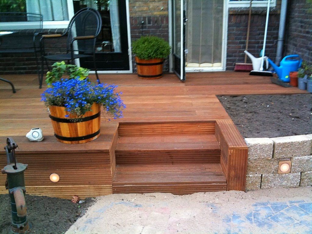 Houten terras met trap, bloempotten en een kleine tuin met bakstenen rand.