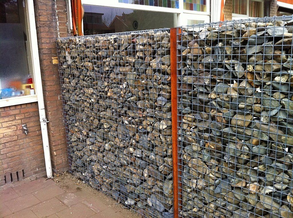 Een met stenen gevulde gabionmuur staat naast een bakstenen gebouw met een raam.
