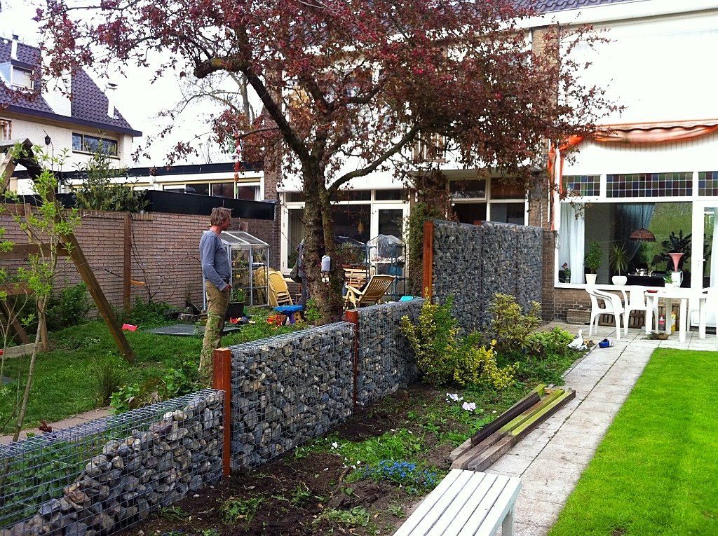 Een persoon staat bij een schanskorfmuur in een achtertuin met een huis, een boom en een gazon.