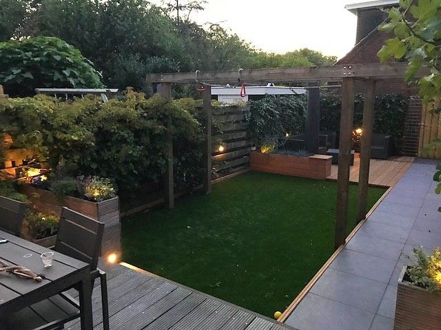 Achtertuin met kunstgras, houten terras, pergola en sfeerverlichting.
