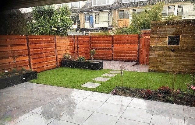Een achtertuin op een regenachtige dag met een houten schutting, gras, stapstenen en een modern terras.