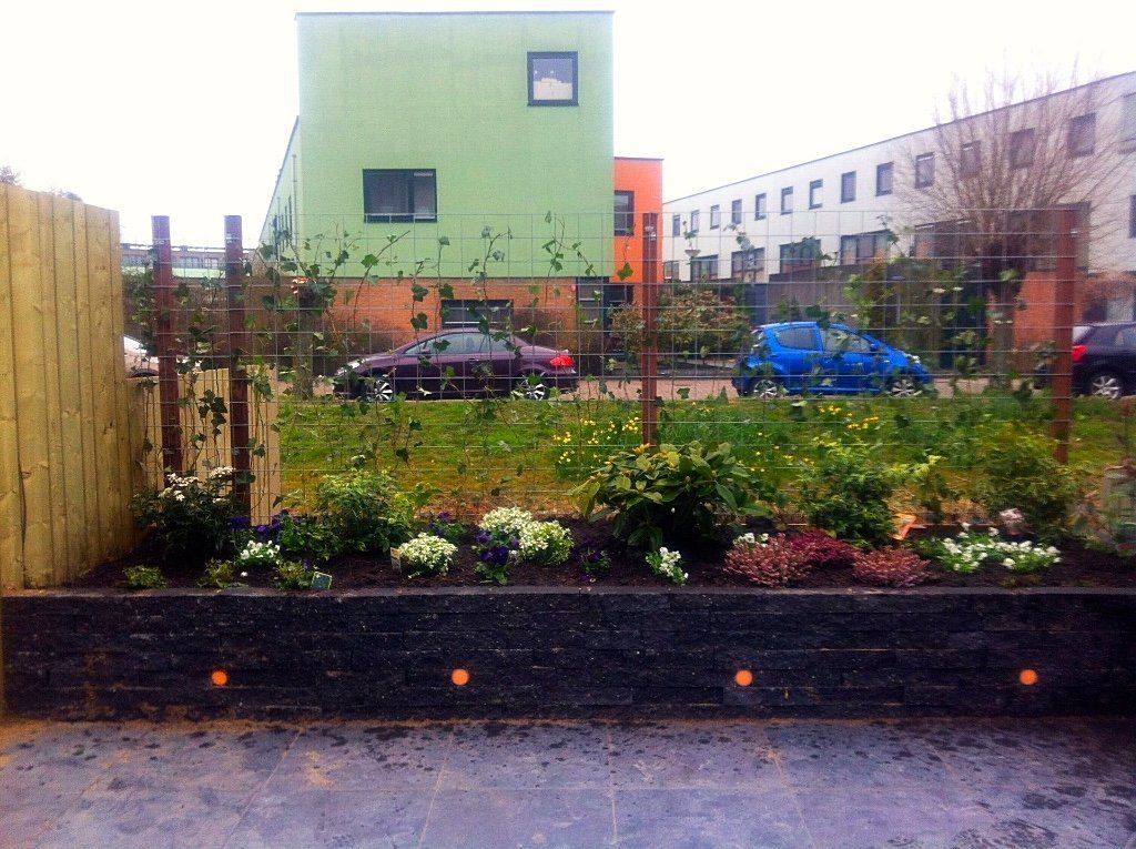 Bloementuin met draadhek, voor een kleurrijk appartementencomplex. Auto's geparkeerd langs een straat.