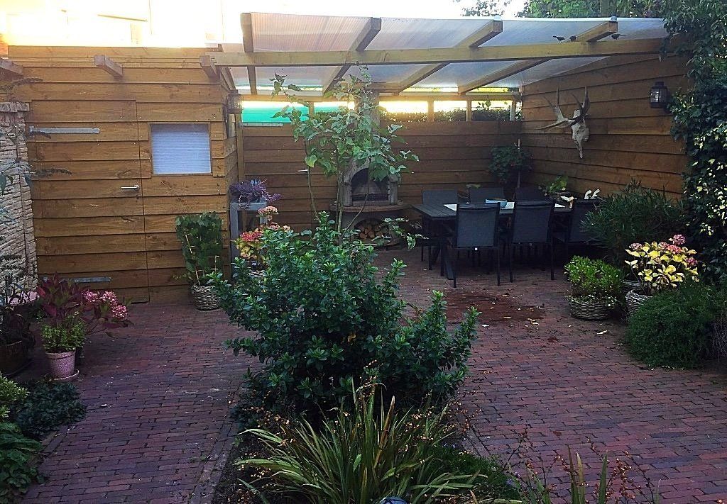 Een terras in de achtertuin met een houten schutting, een eettafel en diverse planten. Een pergola overdekt het zitgedeelte.