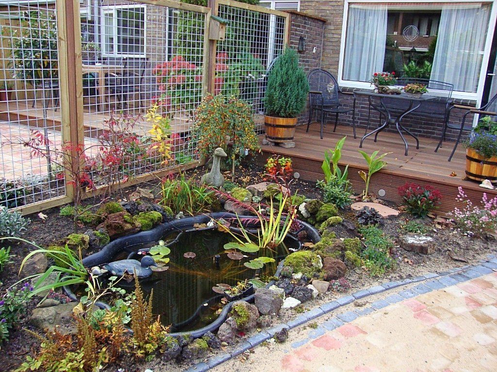 Een tuin met een vijver, planten en een houten terras met tuinmeubilair.