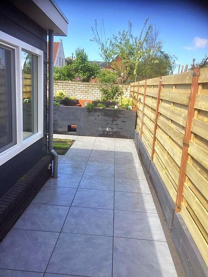 Terras met grijze tegels, houten schutting, gevel van het huis en kleine tuin. Een heldere zonnige dag.