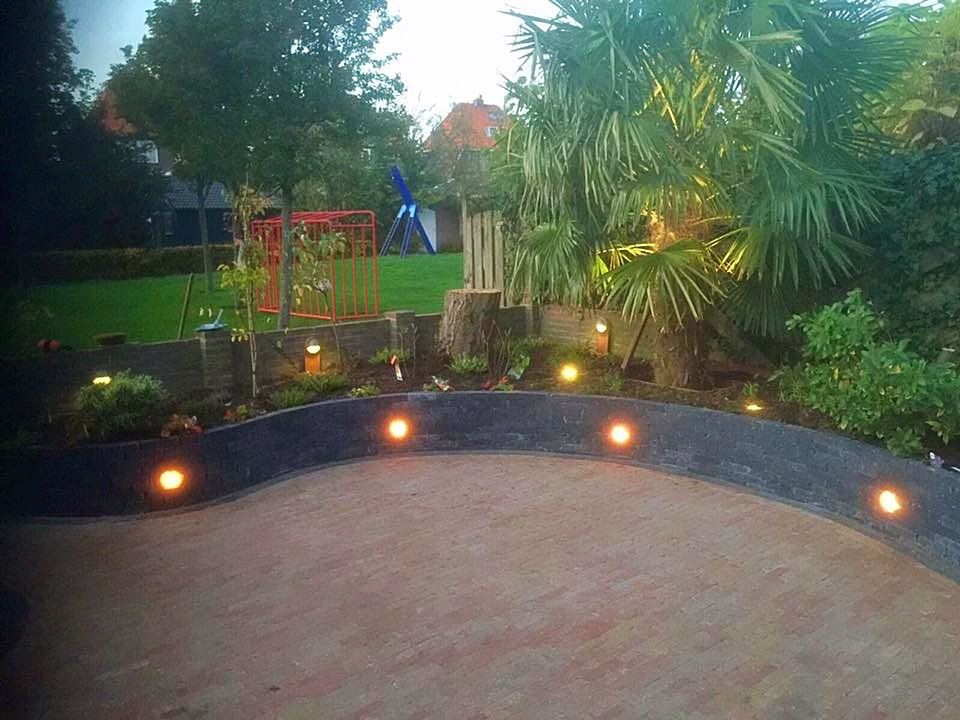 Een gebogen terras met verlichting op grondniveau, dat uitkomt op een aangelegde tuin met bomen, gras en een speeltuin.