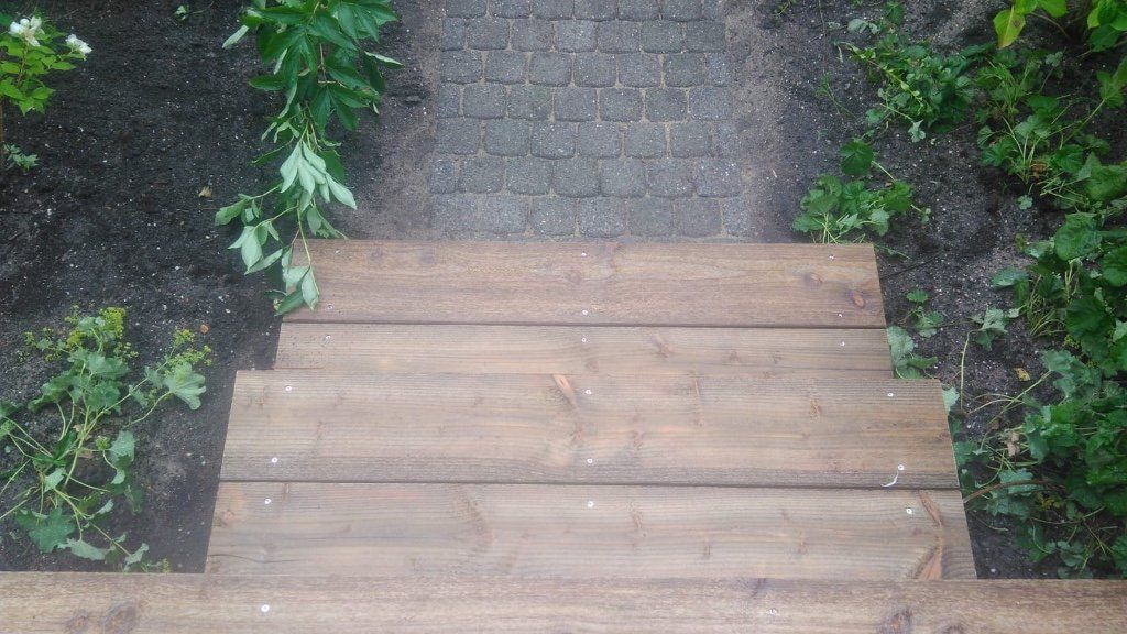 Houten trappen die leiden naar een bakstenen pad, omgeven door groene planten in een buitenomgeving.